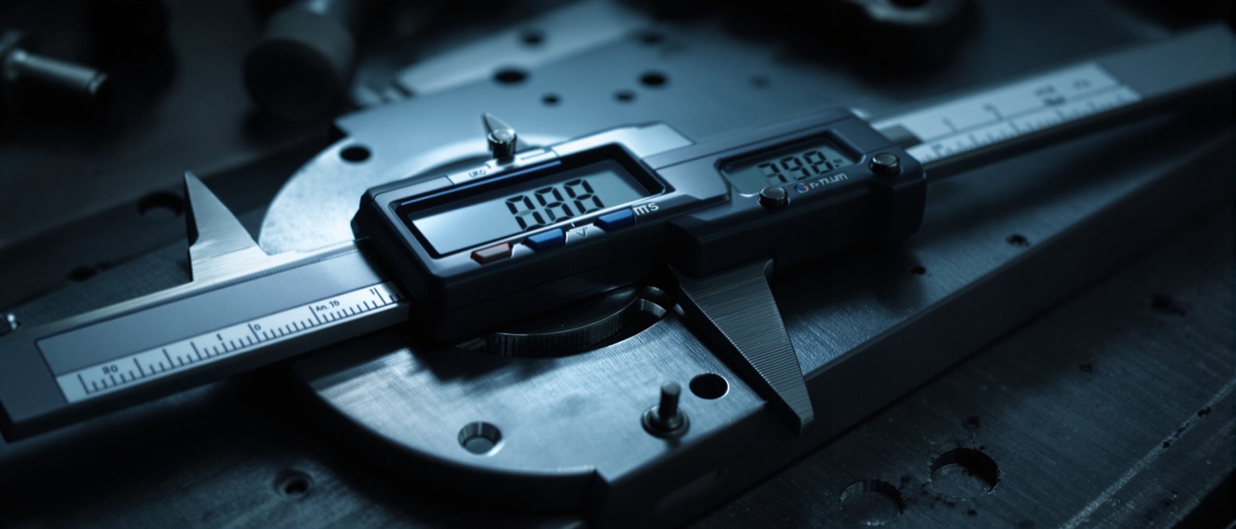 Digital calipers and a micrometer resting on a machined aluminium part on a steel bench.